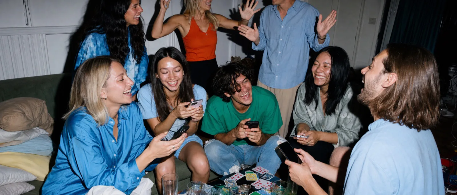 A living room full of friends on and around a couch, all laughing, phones in hand, playing cards and drinks scattered across the table in front of them.